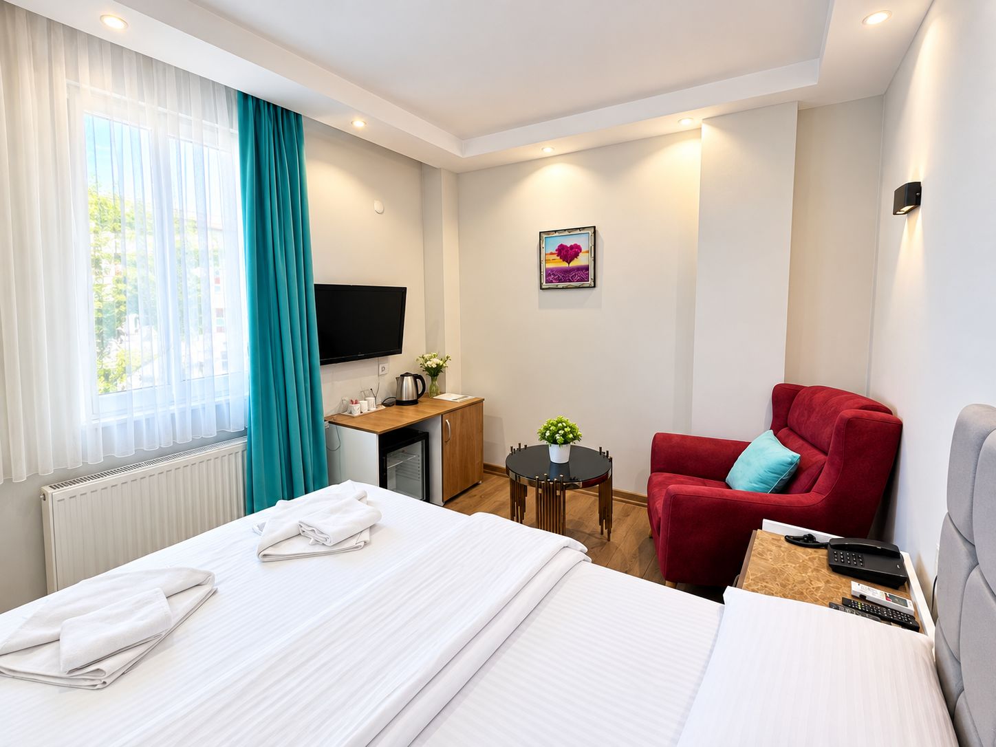 A hotel room with a bed, a desk, a red armchair, a television, and a window with curtains.