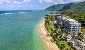 Luftaufnahme eines Hotels direkt am Strand mit Pool, Strand und Bergen im Hintergrund.