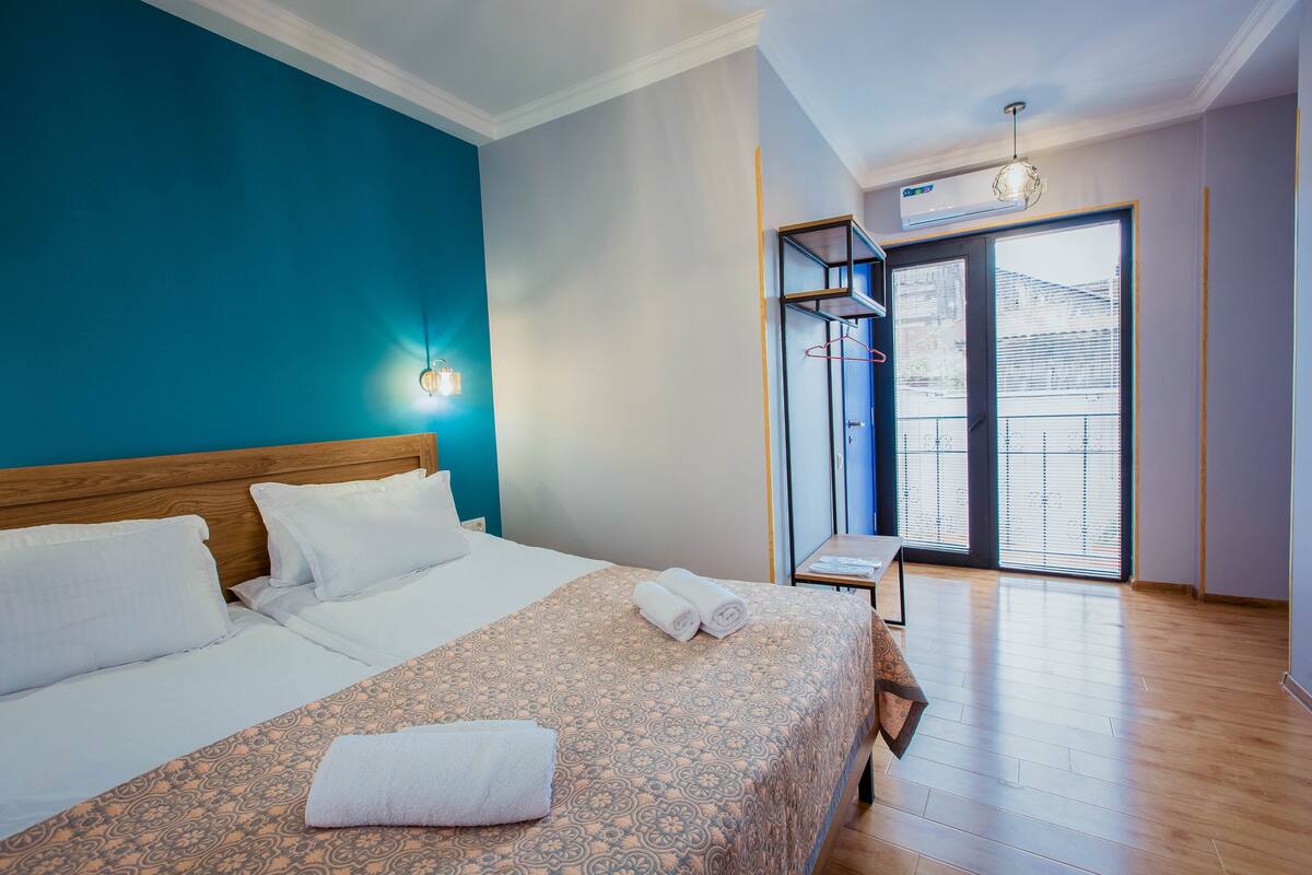 A hotel room with a bed, a nightstand, a window with blinds, and a ceiling-mounted air conditioner.