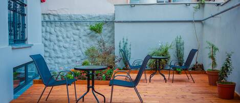 An outdoor patio with wooden flooring, metal chairs, and tables. There are potted plants and a small garden area.