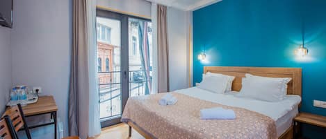 A hotel room with a bed, a desk, a chair, a balcony with a view of buildings, and a blue wall.