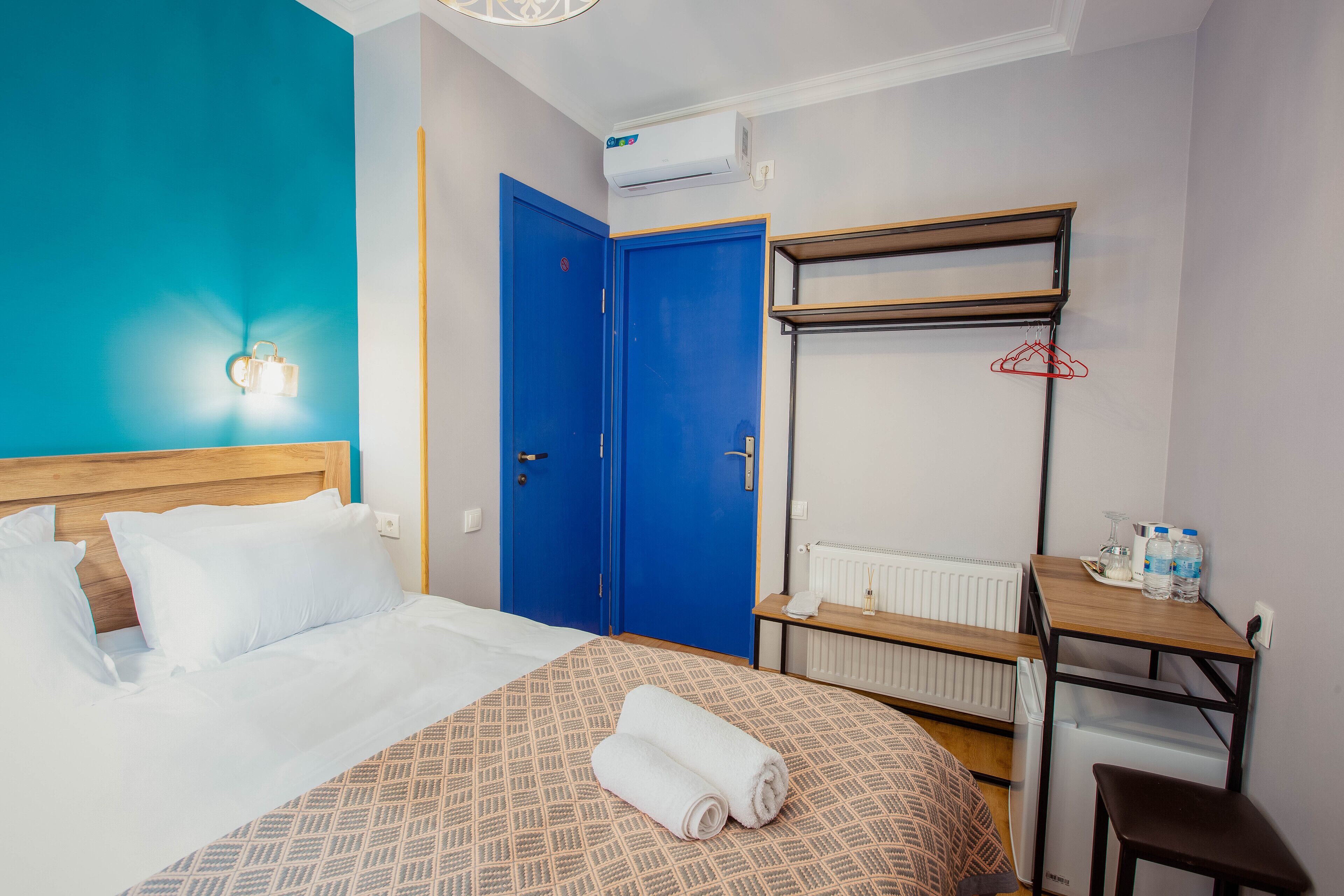 A hotel room with a bed, a blue door, a radiator, a small table, and a wall-mounted shelf.