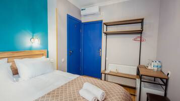 Una habitación de hotel con cama, una puerta azul, un radiador, una mesita y un estante en la pared.