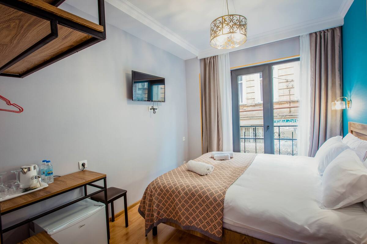 A hotel room with a bed, a desk with a computer, a TV, and a window with curtains.