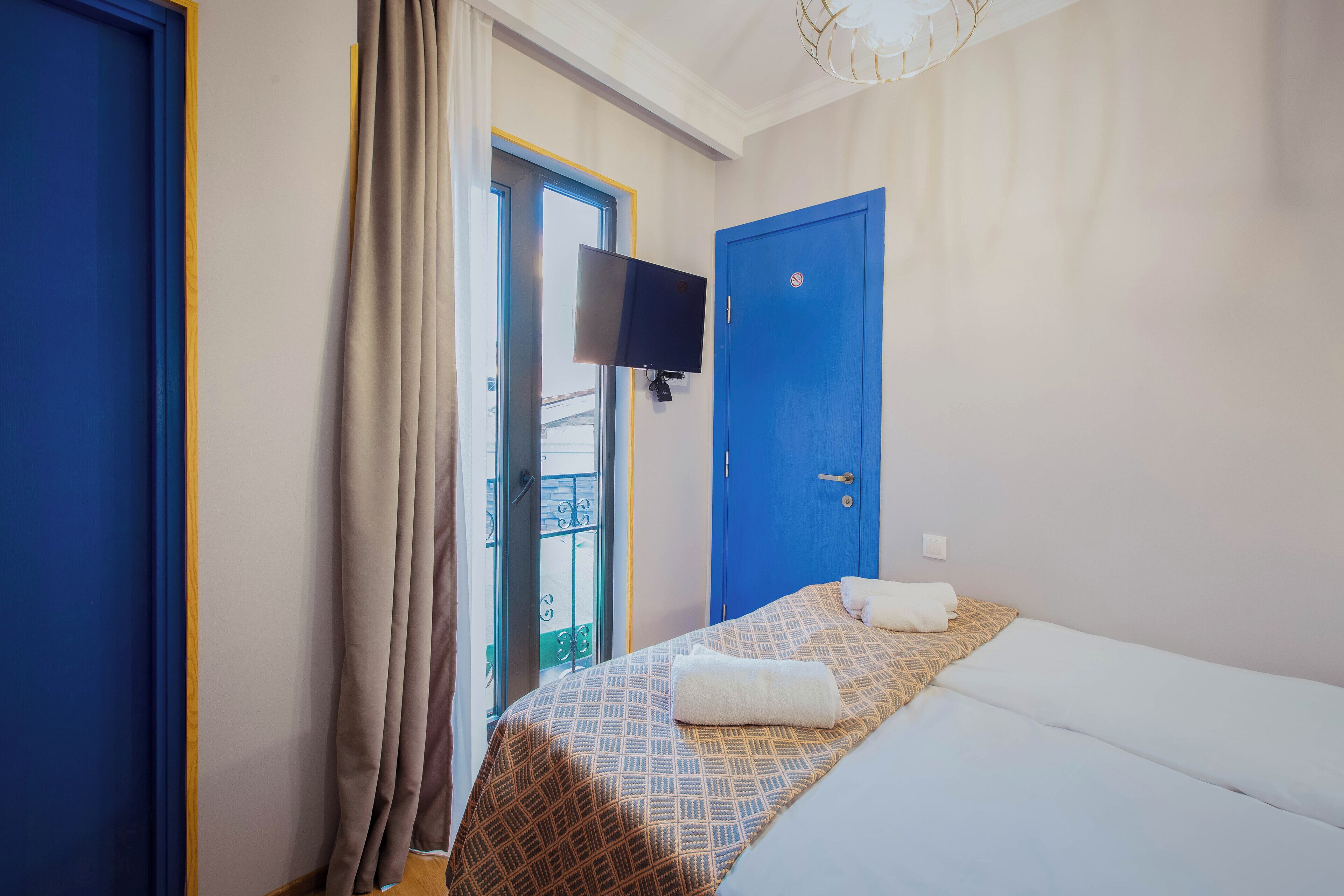 A hotel room with a bed, a blue door, a TV mounted on the wall, and a balcony with curtains.