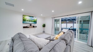 Una sala de estar moderna con un sofá gris, un televisor de pantalla plana montado en la pared y un ventanal corredero que da a un área exterior.