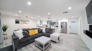Una sala de estar moderna con un sofá seccional gris, un otomano blanco y un comedor con una mesa pequeña y sillas.