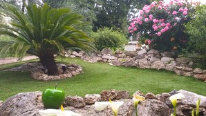 A garden with a palm tree, a rock with a green apple, white flowers, and pink flowers. - Superbe 3P dans villa avec vue mer et collines (Carros)