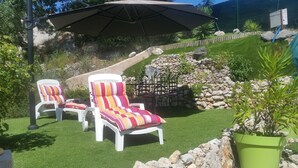 A patio area with two white lounge chairs with striped cushions, a large umbrella, and a stone wall with greenery. - Superbe 3P dans villa avec vue mer et collines (Carros)