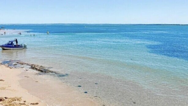 Una playa con aguas transparentes y poco profundas, un bote en la orilla y gente en el agua.