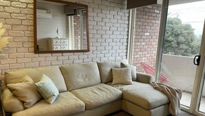Un sofá seccional color beige con varios cojines, una alfombra estampada, un espejo y una pared de ladrillo.