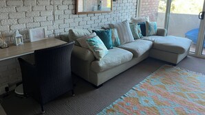 Una sala de estar con pared de ladrillo, un ventanal grande, un sofá con cojines, un sillón de mimbre y una mesita con una vela y un marco de fotos.