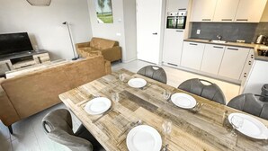 Ein moderner Essbereich mit einem Holztisch, gedeckt für vier Personen, umgeben von grauen Sitzstühlen. Im Hintergrund sind eine Couch und ein Fernseher zu sehen.