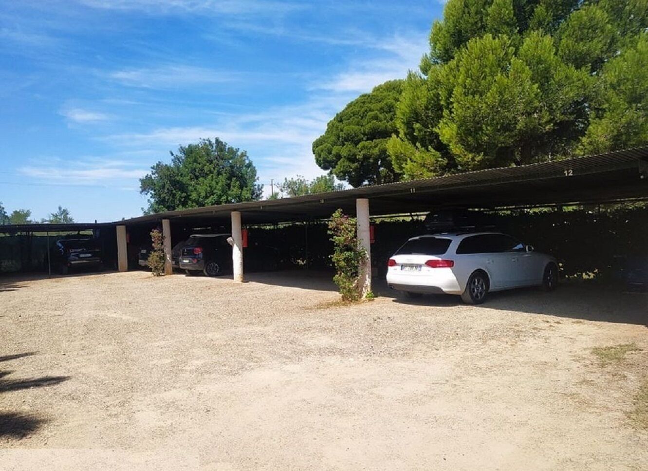 Un área de estacionamiento con un marquesina cubierta, un coche blanco estacionado bajo la protección y un suelo de grava.