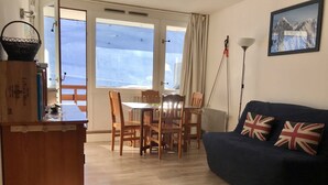 Olohuoneessa on tummansininen sohva, puiset tuolit, puinen pöytä ja ikkuna, jossa on verhot.