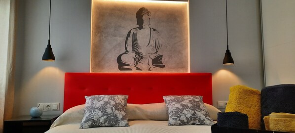 A bedroom with a red headboard, two floral pillows, a bed with a textured blanket, and a wall-mounted artwork of a seated figure.