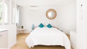 A bright, white room with a bed, a small round mirror, and a window with curtains.