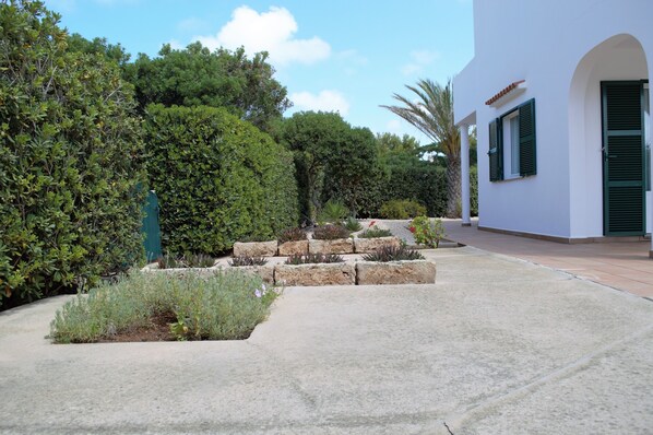 A pathway leading to a white building with green shutters, surrounded by lush greenery and a small garden area.