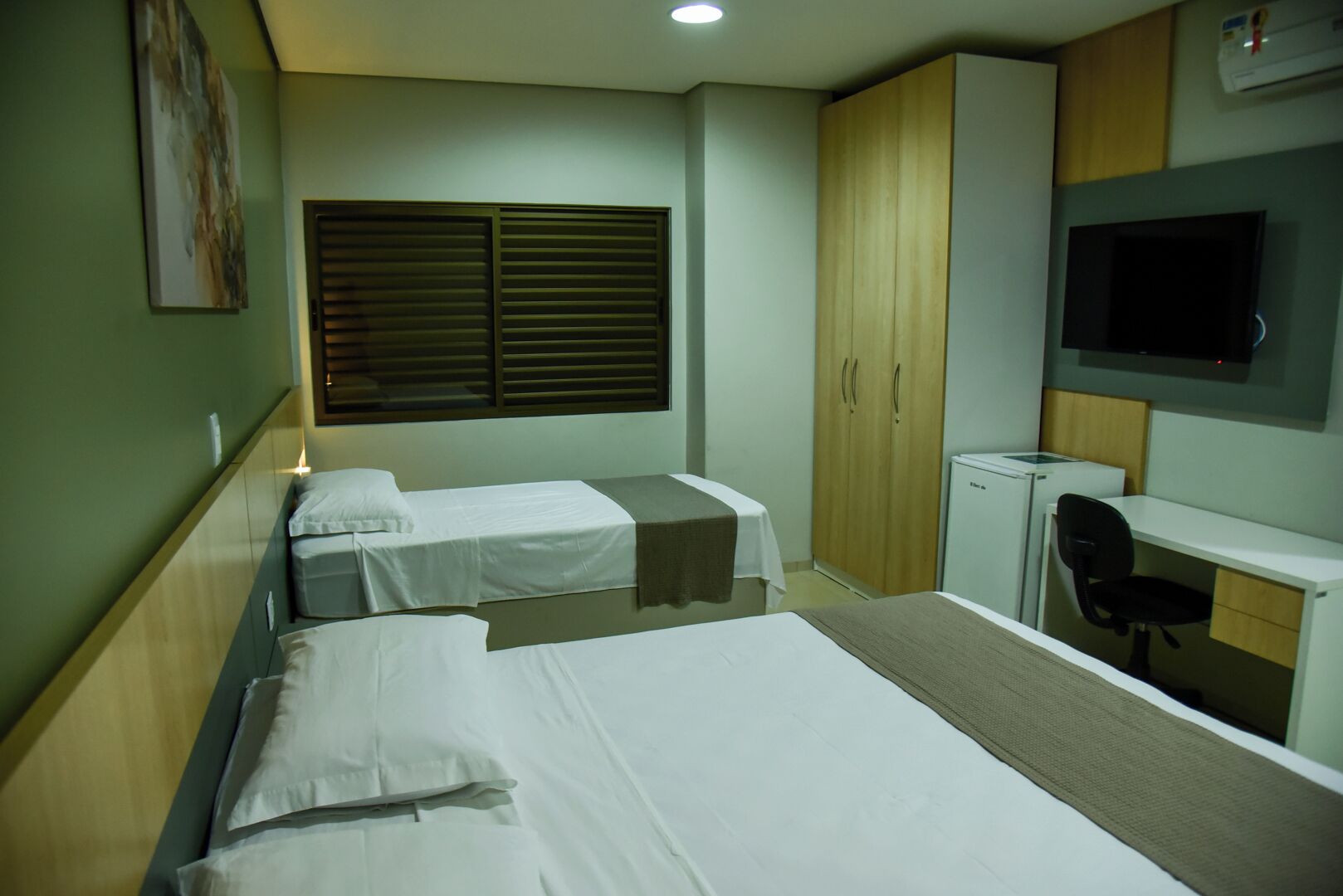 A hotel room with two single beds, a desk with a chair, a wardrobe, and a television on the wall.
