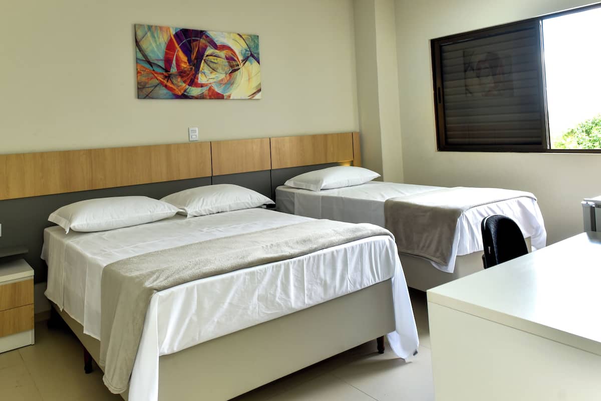 A hotel room with two beds, a wooden headboard, a bedside table with a lamp, a painting on the wall, and a window with blinds.