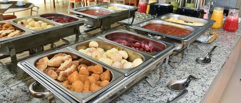 Un bufé con diversos alimentos en recipientes metálicos, como pan, pastelería y diferentes tipos de carne.