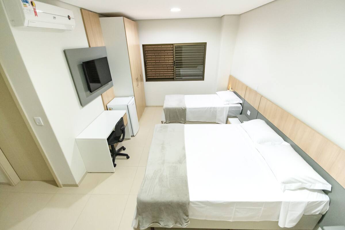 A hotel room with two beds, a desk, a TV, and a window with blinds.