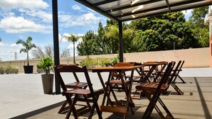 Un patio al aire libre con mesas y sillas de madera, rodeado de plantas en macetas y árboles.