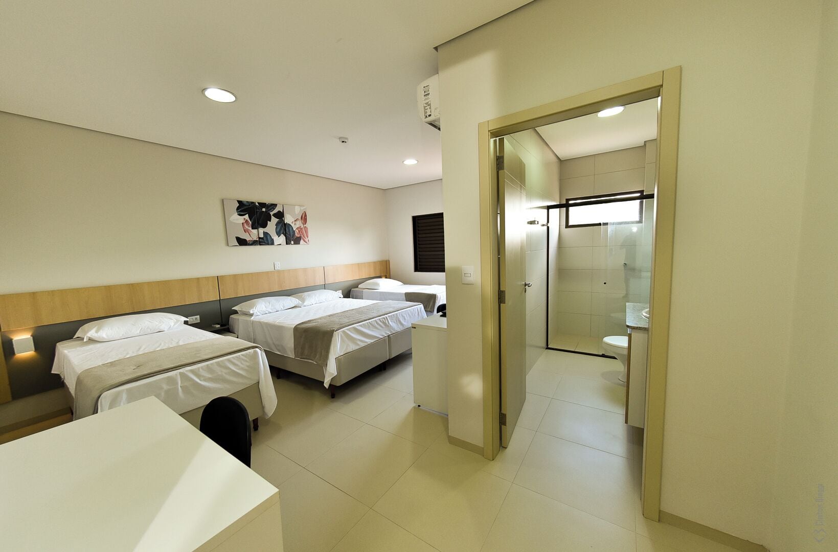 Quarto de hotel com duas camas, uma mesinha e um banheiro visível através de uma porta aberta.