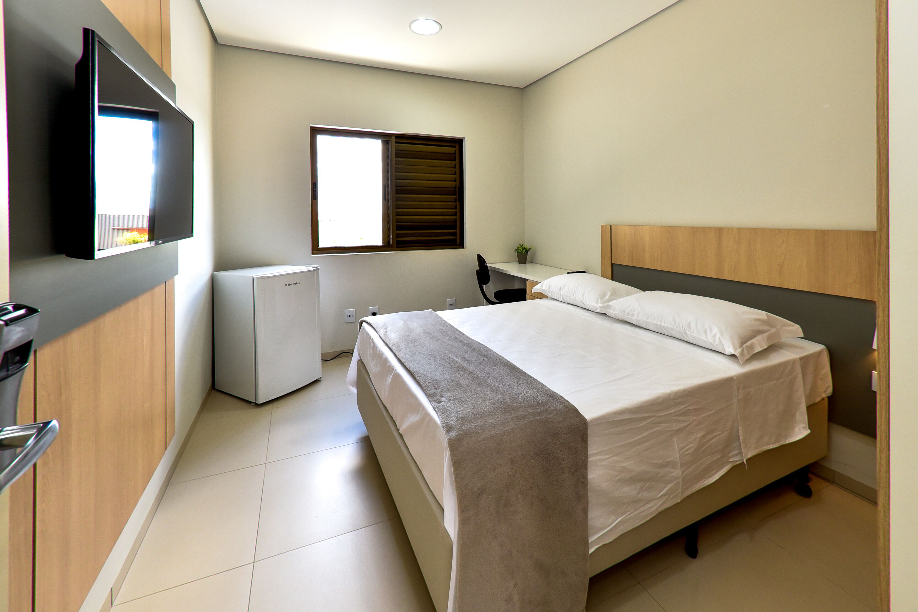 A bedroom with a bed, a desk, a chair, a TV, and a window.