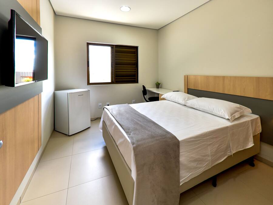 A bedroom with a bed, a desk, a chair, a TV, and a window.