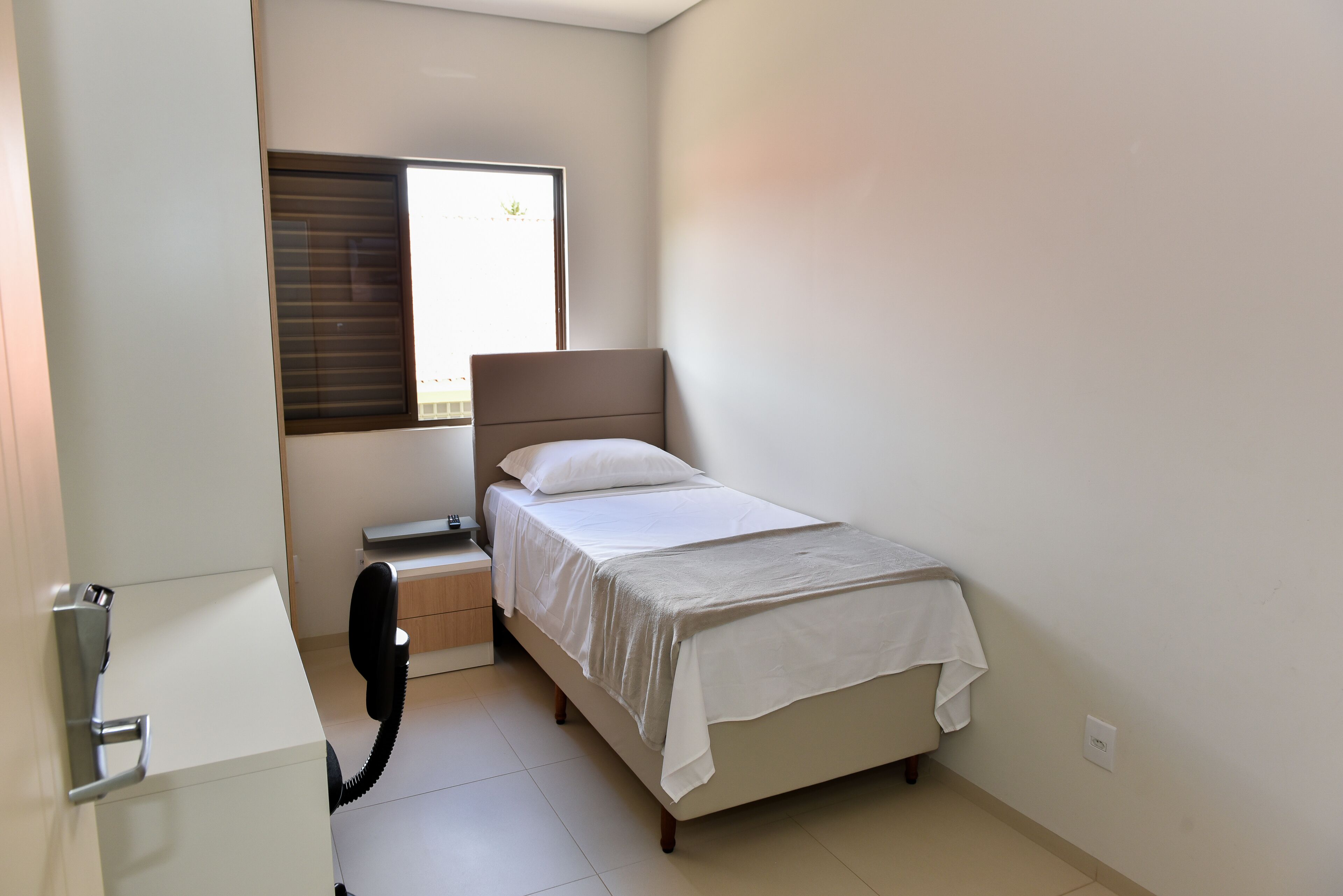 Une petite chambre propre, avec un lit, une table de chevet, un téléphone et une fenêtre avec des stores.