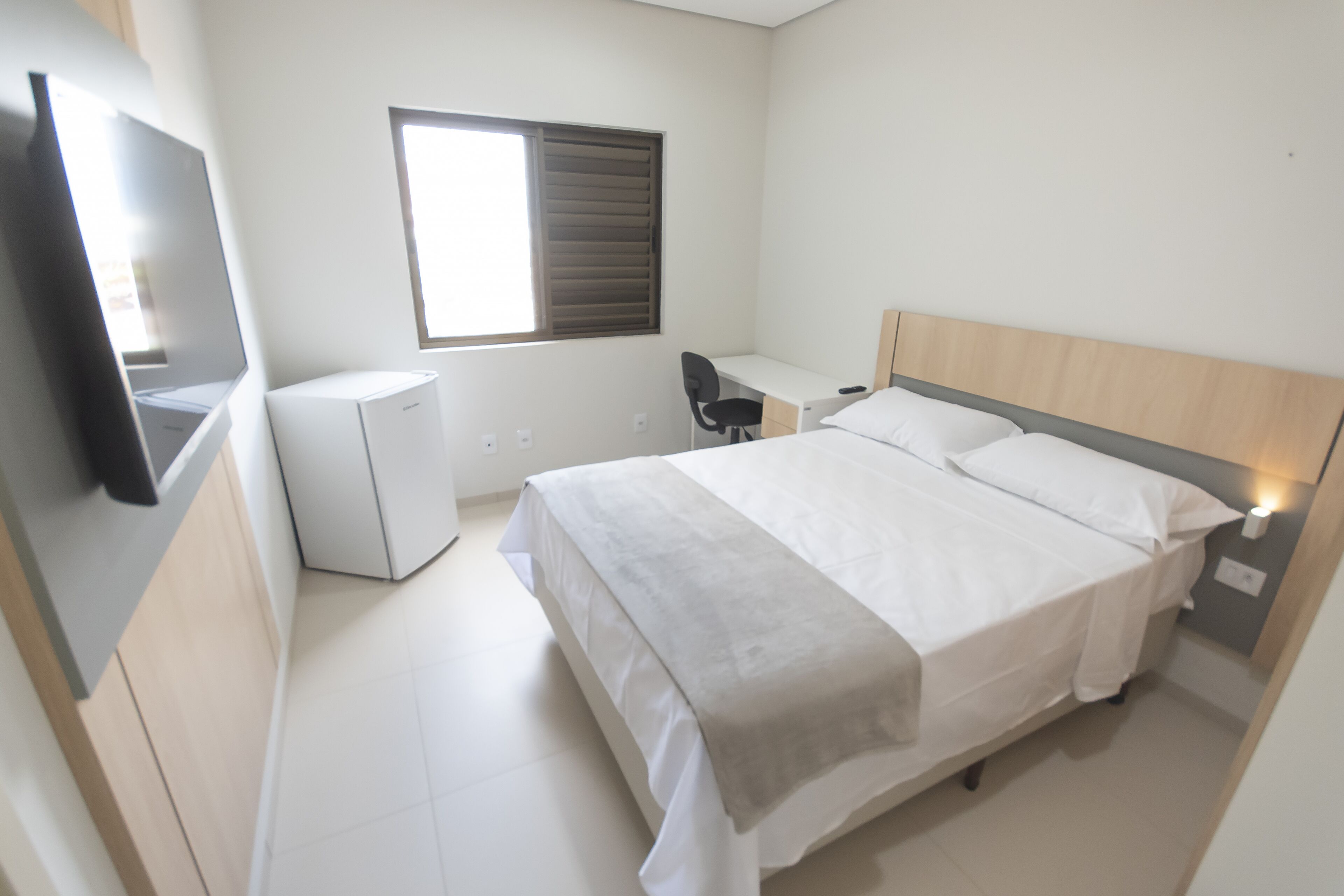 A bedroom with a bed, a desk, a chair, a TV, and a window.