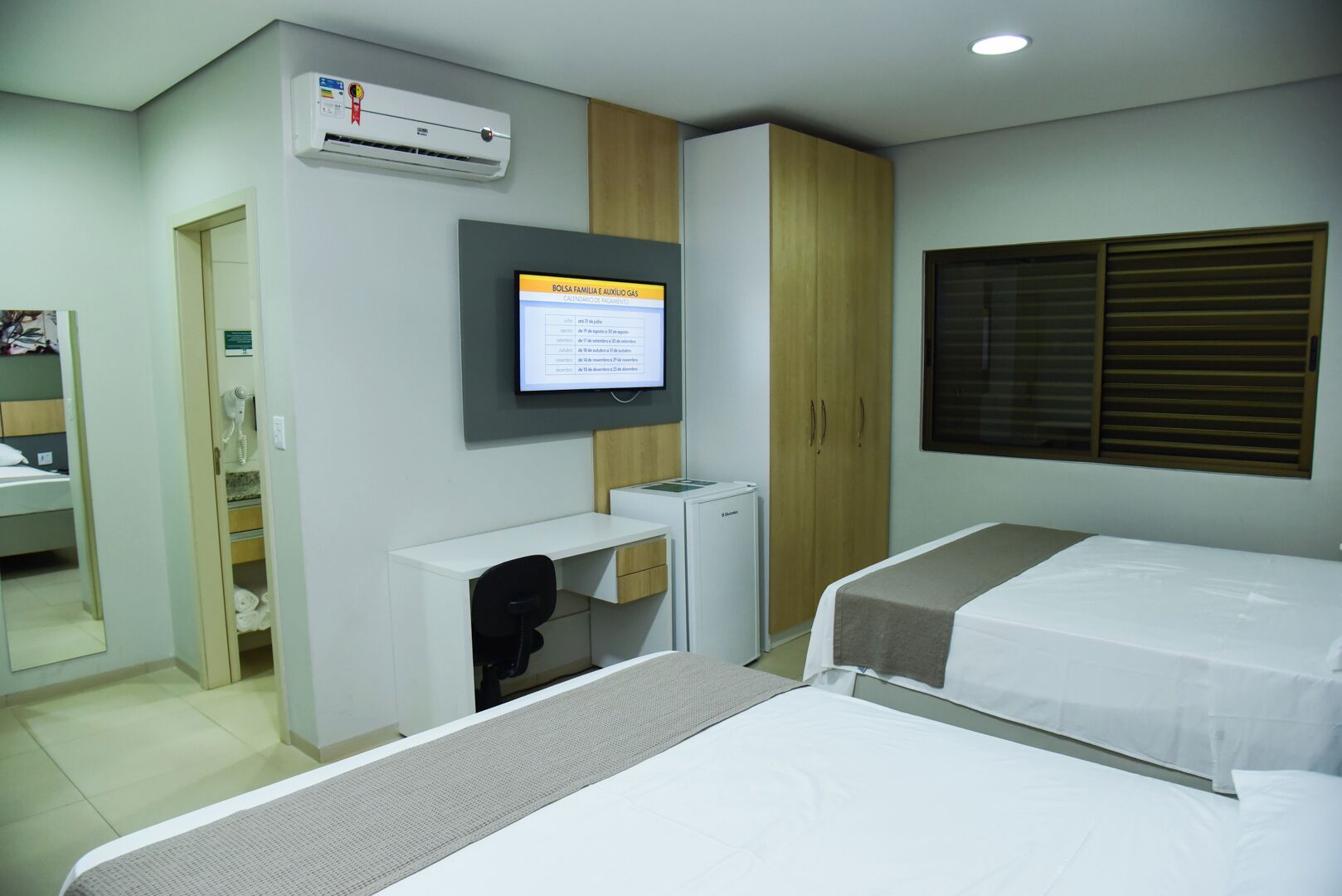 A hotel room with two beds, a desk, a wardrobe, an air conditioner, and a television displaying a menu.