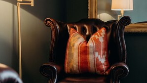 Een donkere leren fauteuil met een kussen met een patroon en een plaid, geplaatst voor een vloerlamp en een spiegel op een tapijtvloer.