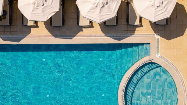 A swimming pool with white umbrellas and lounge chairs, surrounded by a tiled deck and yellow flowering plants. - Nora Suit Hotel (Manavgat)