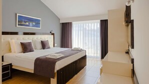 A hotel room with a bed, a TV, a window with curtains, and a painting on the wall. - Nora Suit Hotel (Manavgat)