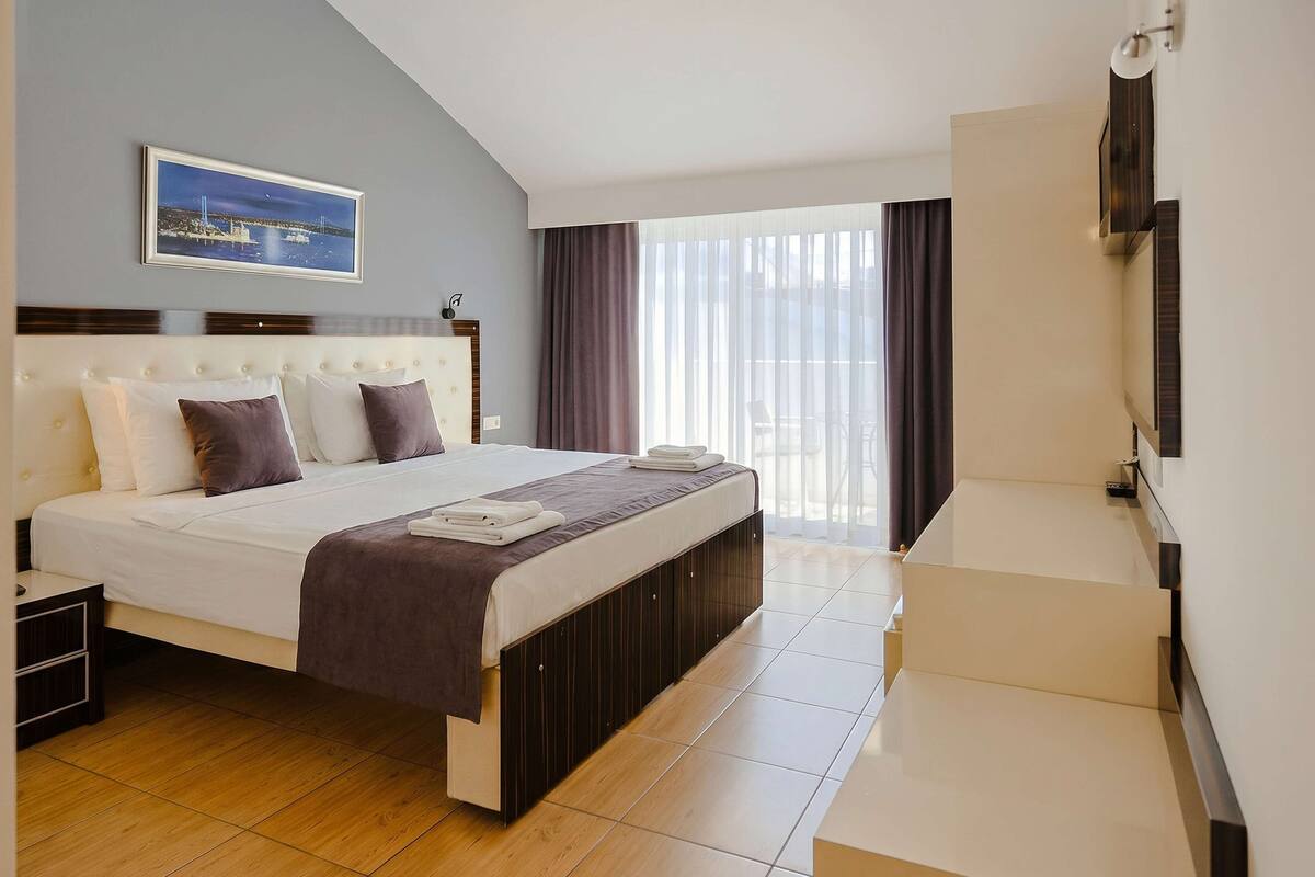 A hotel room with a bed, a TV, a window with curtains, and a painting on the wall.