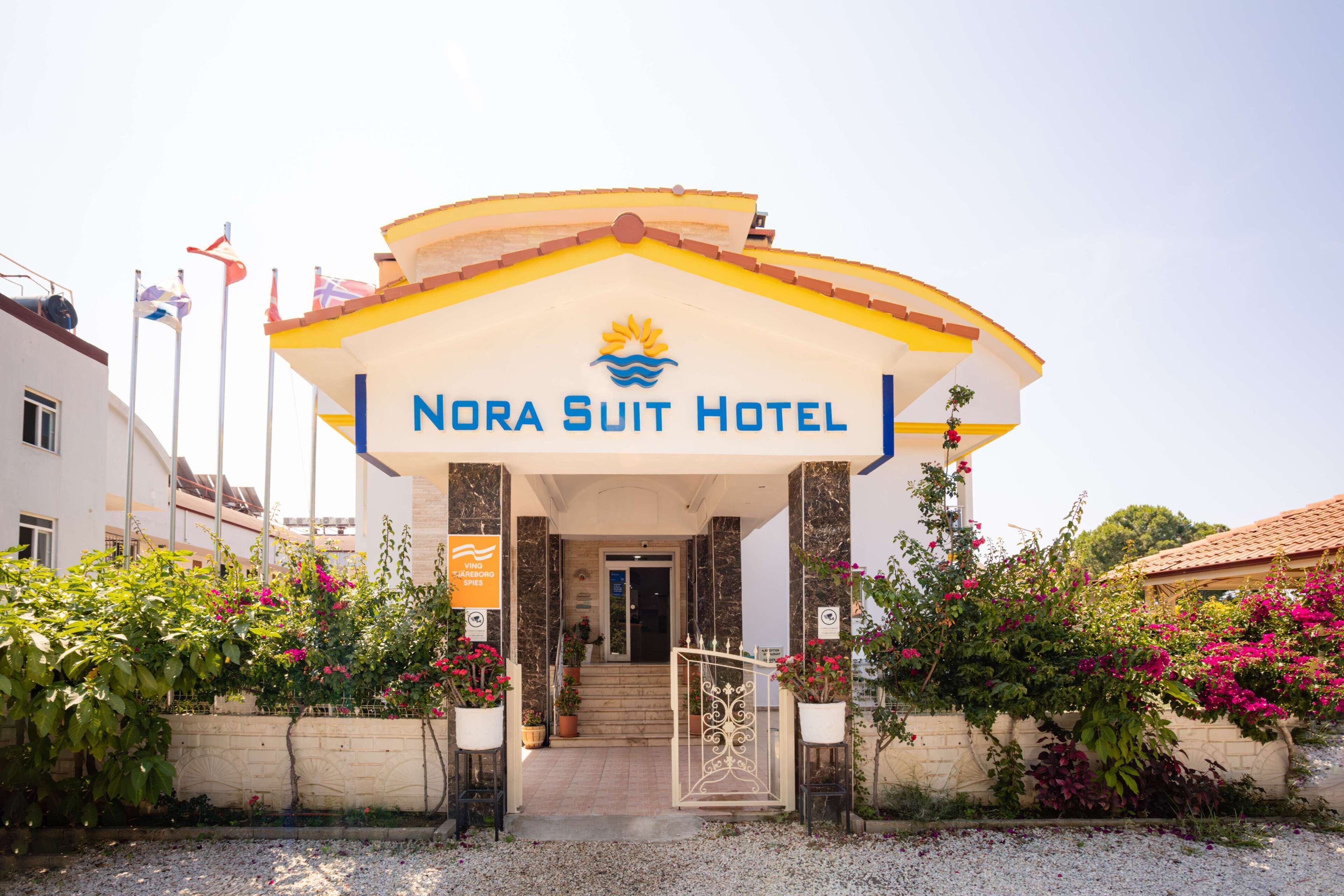 Exterior del Hotel Nora Suit, con una entrada destacada, un cartel y follaje circundante.