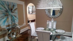 A dining area with a glass table, white chairs, a large abstract painting, a mirror, and a chandelier. - The Le Chic Bungalow (Decatur)