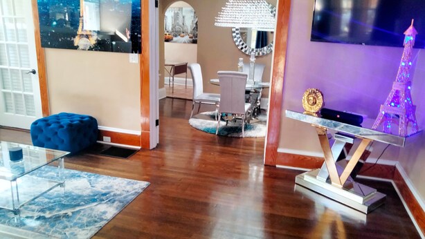 A spacious living room with wooden flooring, a glass coffee table, a blue ottoman, a chandelier, and a TV mounted on the wall. - The Le Chic Bungalow (Decatur)