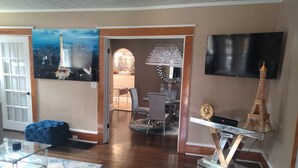 A modern living room with a wooden floor, a glass coffee table, a blue ottoman, a dining area with a chandelier, and a TV mounted on the wall. - The Le Chic Bungalow (Decatur)