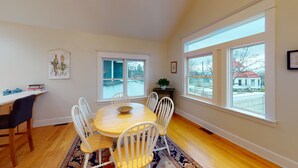A dining room with a wooden table and white chairs, large windows with a view of a snowy landscape, and a small plant on a side table. - Wonderful Main Street Location, next to the HS Historical Society! (Harbor Springs)