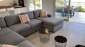 A modern living room with a grey sectional sofa, two black wireframe side tables, and a glass sliding door leading to a balcony with outdoor furniture. - spacious appartment facing the sea with private garden jaccuzi and plancha grill (Saint Martin)