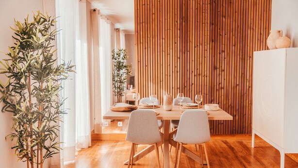 Un comedor con una mesa de madera dispuesta para cuatro personas, rodeada de sillas de colores claros. Se observa una planta en maceta a la izquierda.