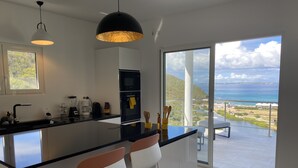 Una cocina moderna con una isla grande, un rincón comedor con dos sillas y vista al océano.