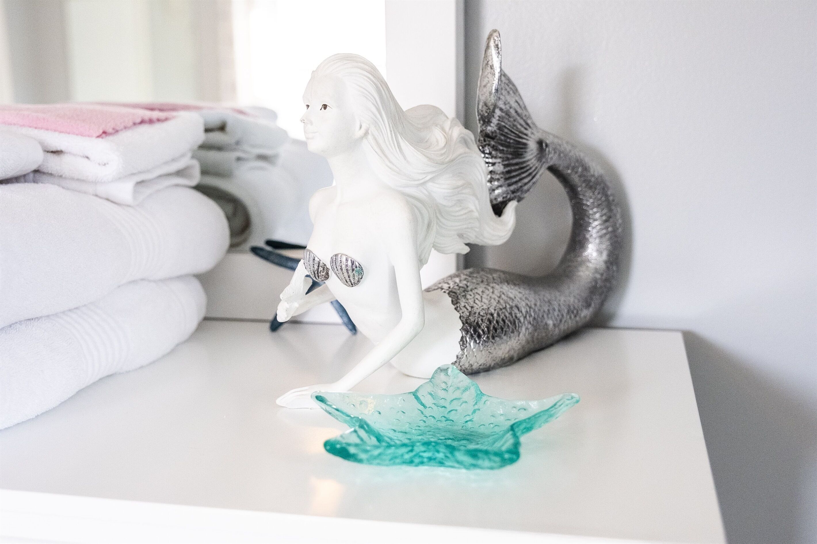 A white statue of a mermaid with a silver tail, a folded pink towel, and a teal glass bowl on a white surface.