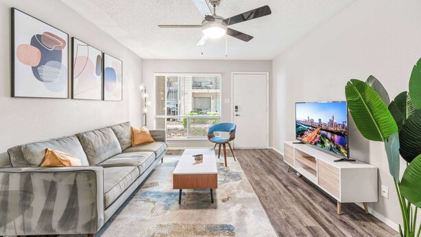 Un salon moderne avec un canapé gris, une table basse, un téléviseur posé sur un meuble blanc et un ventilateur de plafond.
