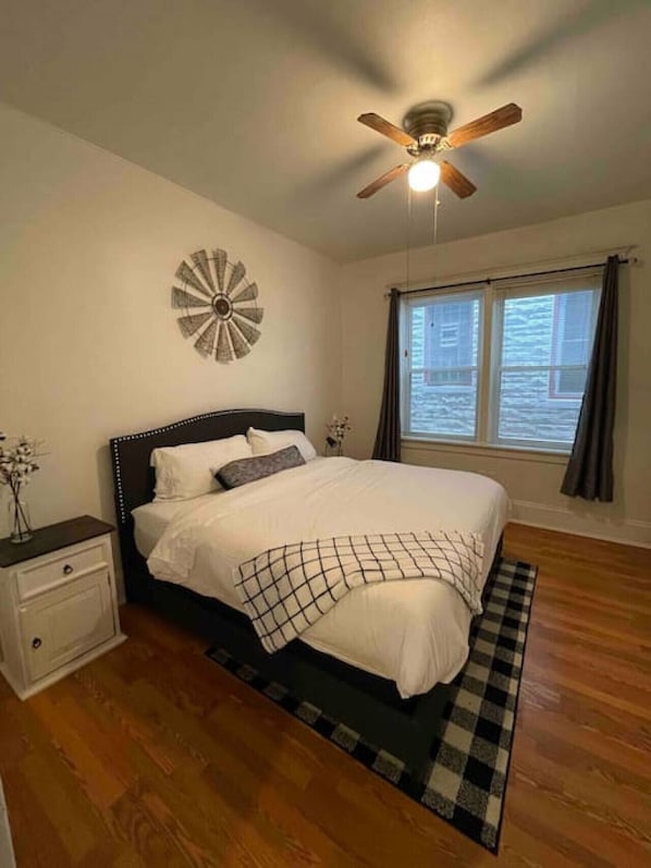 Une chambre à coucher comprenant un lit, une table de chevet, un ventilateur de plafond, une fenêtre avec des rideaux et une horloge murale.