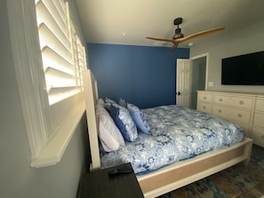 Un dormitorio con cama, ventilador de techo, cómoda y televisor en la pared.