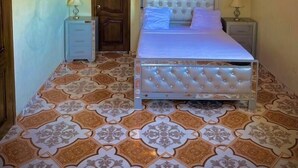 Quarto com cama, duas mesas de cabeceira, porta de madeira e piso com ladrilhos estampados.